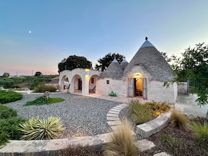Uma casa de pedra com telhado cônico, várias aberturas em arco e um caminho de acesso de cascalho.