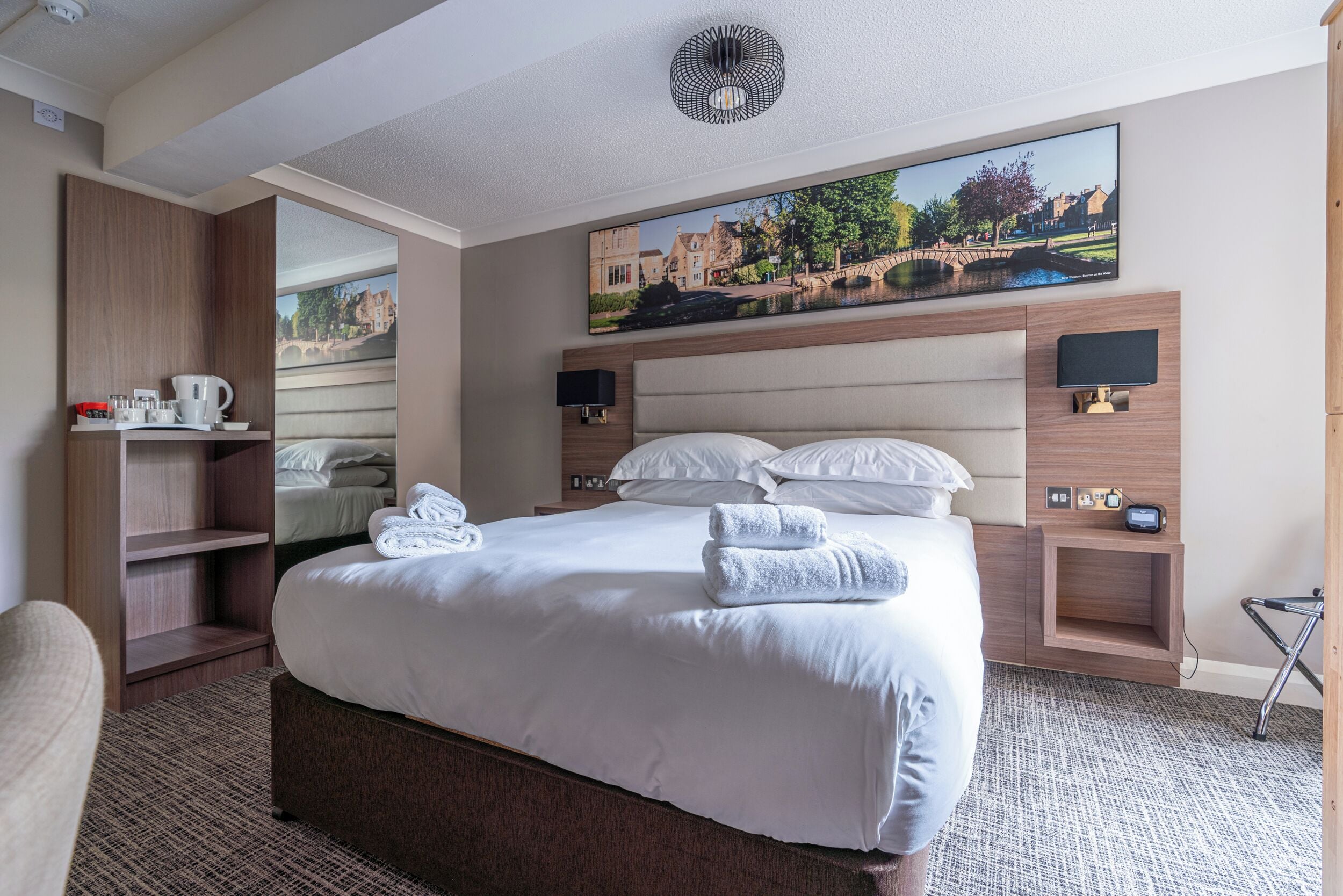 A hotel room with a large bed, a wooden wardrobe, a desk with a chair, a small table with a lamp, and a painting on the wall.