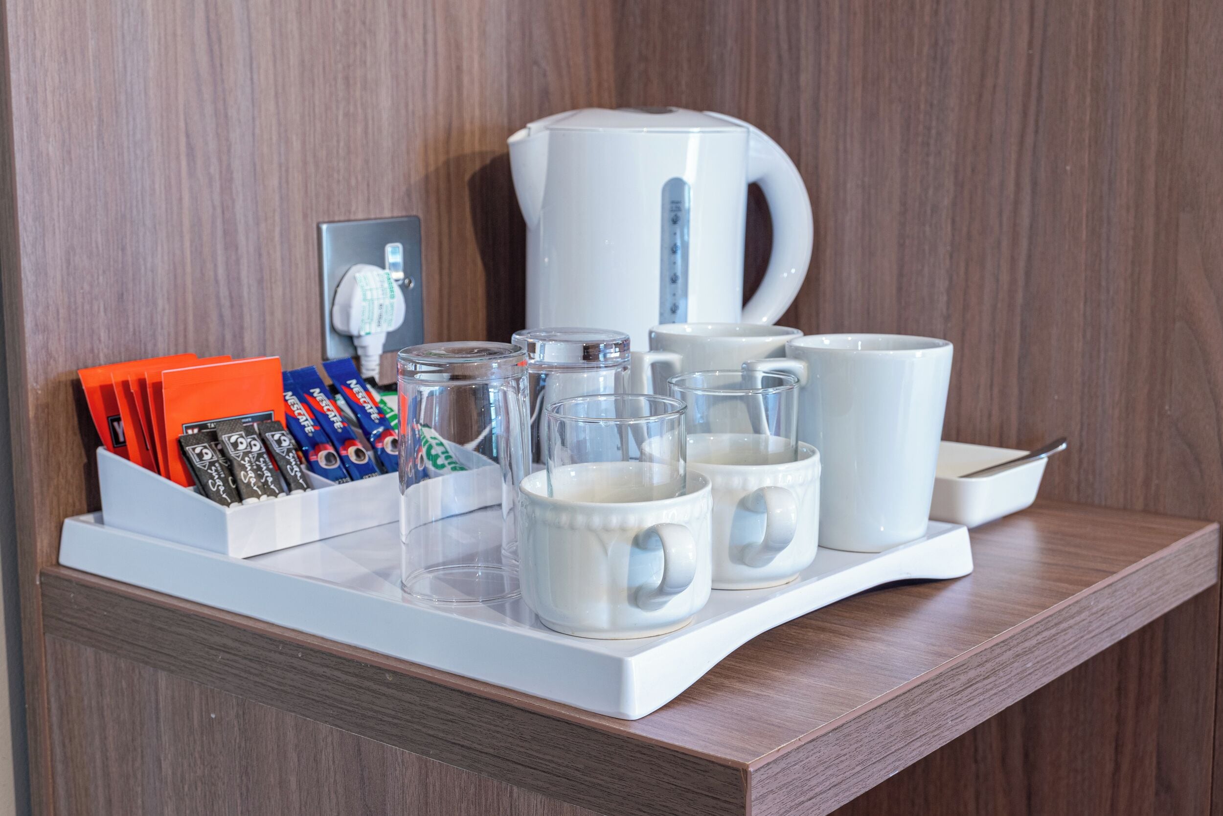 A tray with coffee mugs, a white kettle, and a box of coffee packets on a wooden shelf.