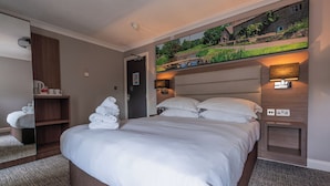 A hotel room with a large bed, a mirror, a flat-screen TV, a bedside table, and a wall-mounted lamp. - Riverside Hotel (Cirencester)