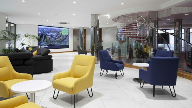 A modern lobby with a large digital display, comfortable seating, and a glass staircase. - Riverside Hotel (Cirencester)