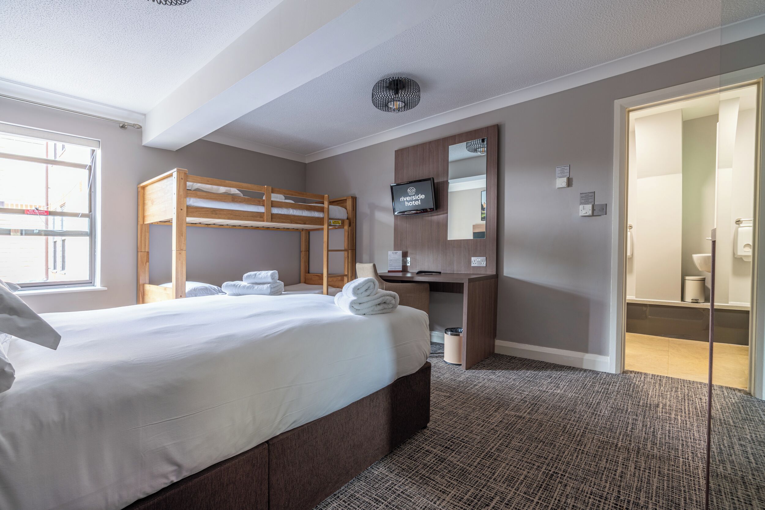 Habitación de hotel con literas, un escritorio, un televisor y un baño visible a través de una puerta abierta.