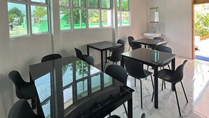 Un'area pranzo moderna con tavoli dal piano in vetro e sedie nere, ampie finestre con vista sul verde, e un soffitto con travi a vista e ventilatori.