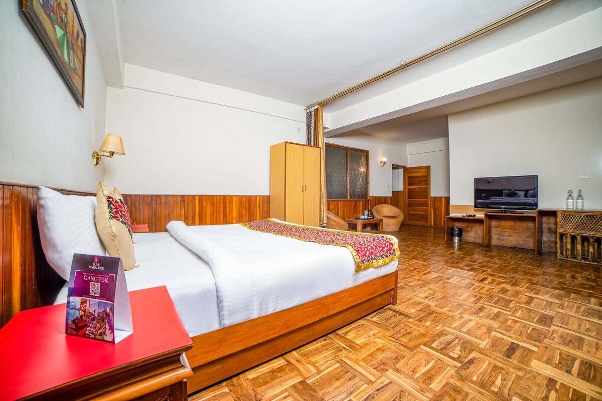 Une chambre d’hôtel avec un plancher en bois, un lit recouvert d’une courtepointe blanche, une table de chevet rouge, une télévision et une armoire en bois.