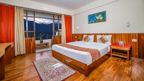 A hotel room with a large bed, wooden headboard, a nightstand, a desk, and a view of mountains through the window. - Hotel Chiminda Pelling (Gyalshing)