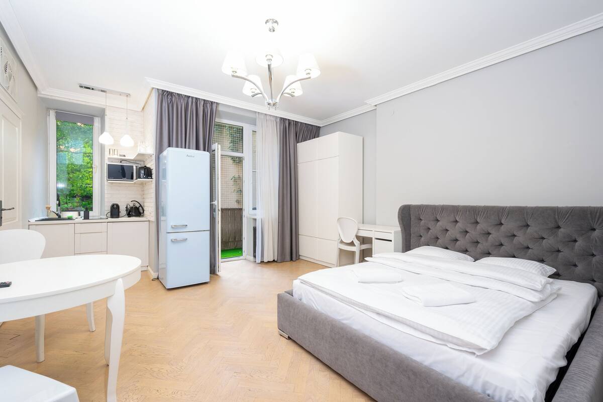 A modern hotel room with a bed, a desk, a chair, a small kitchenette, and a view of the outdoors.