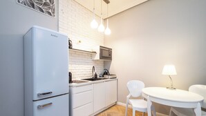 Ett kompakt kök med ett vitt kylskåp, en mikrovågsugn och en diskho. Det finns två vita stolar och ett litet bord med en lampa.