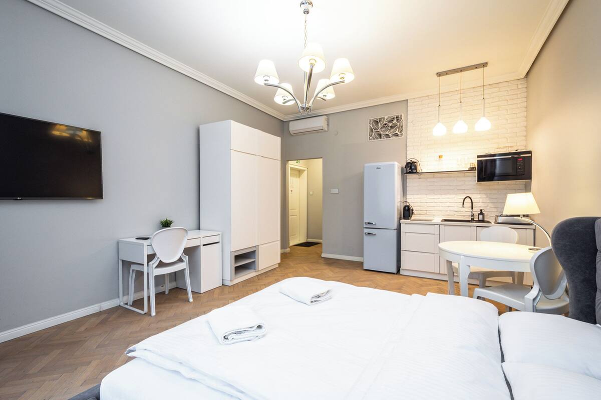 A modern hotel room with a bed, desk, chair, kitchenette, and a TV.