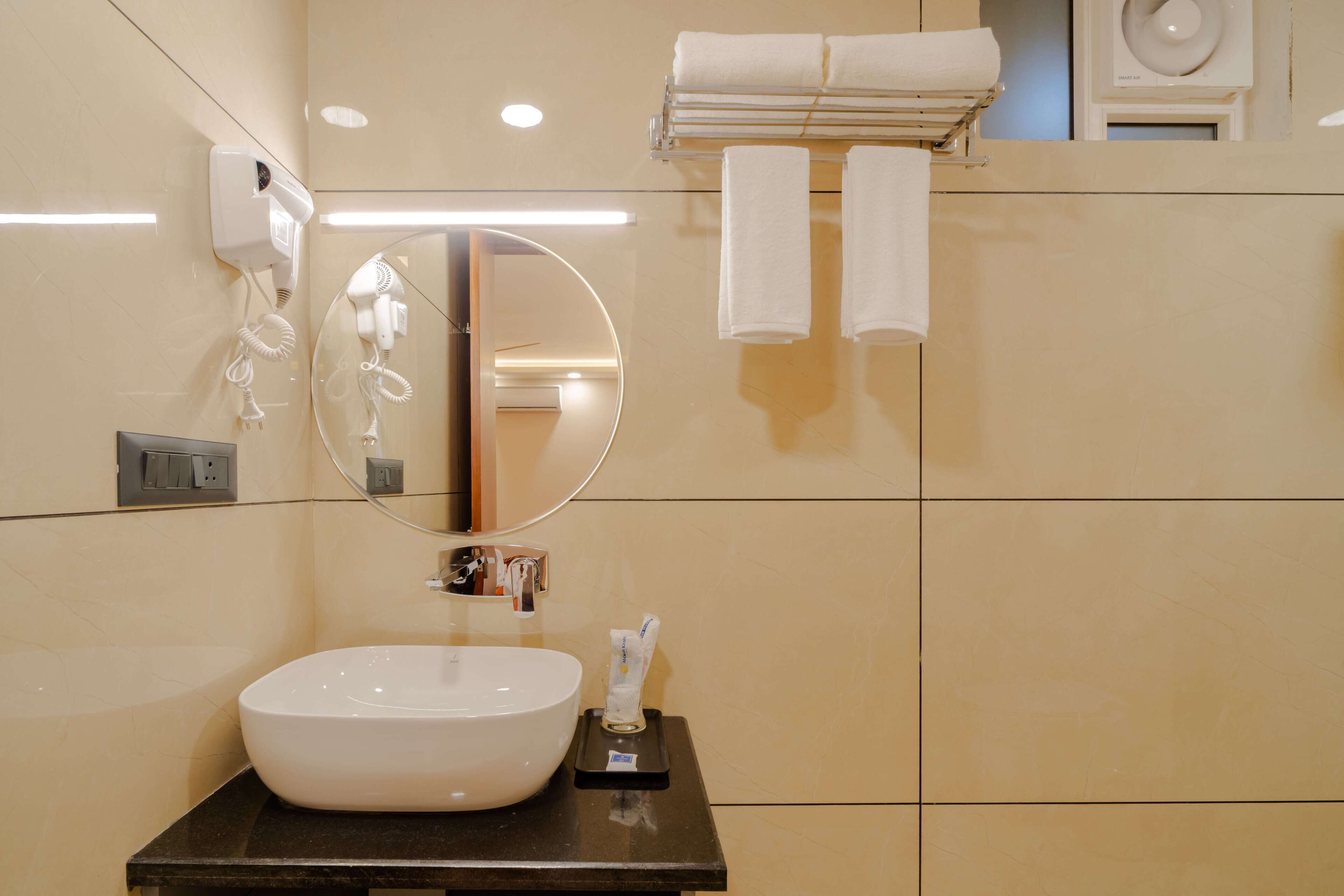 A bathroom with a large oval sink, a round mirror, a towel rack with two towels, and a hairdryer on the wall.