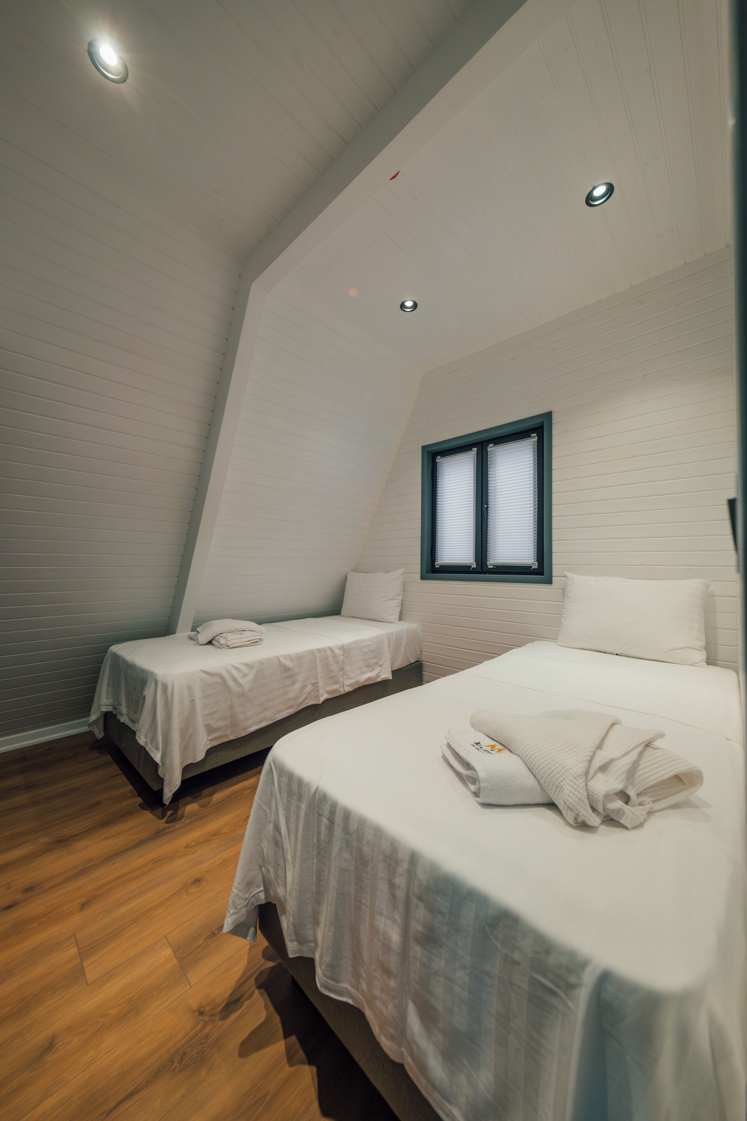 A bedroom with two single beds, a window with blinds, and a sloped ceiling with recessed lighting.