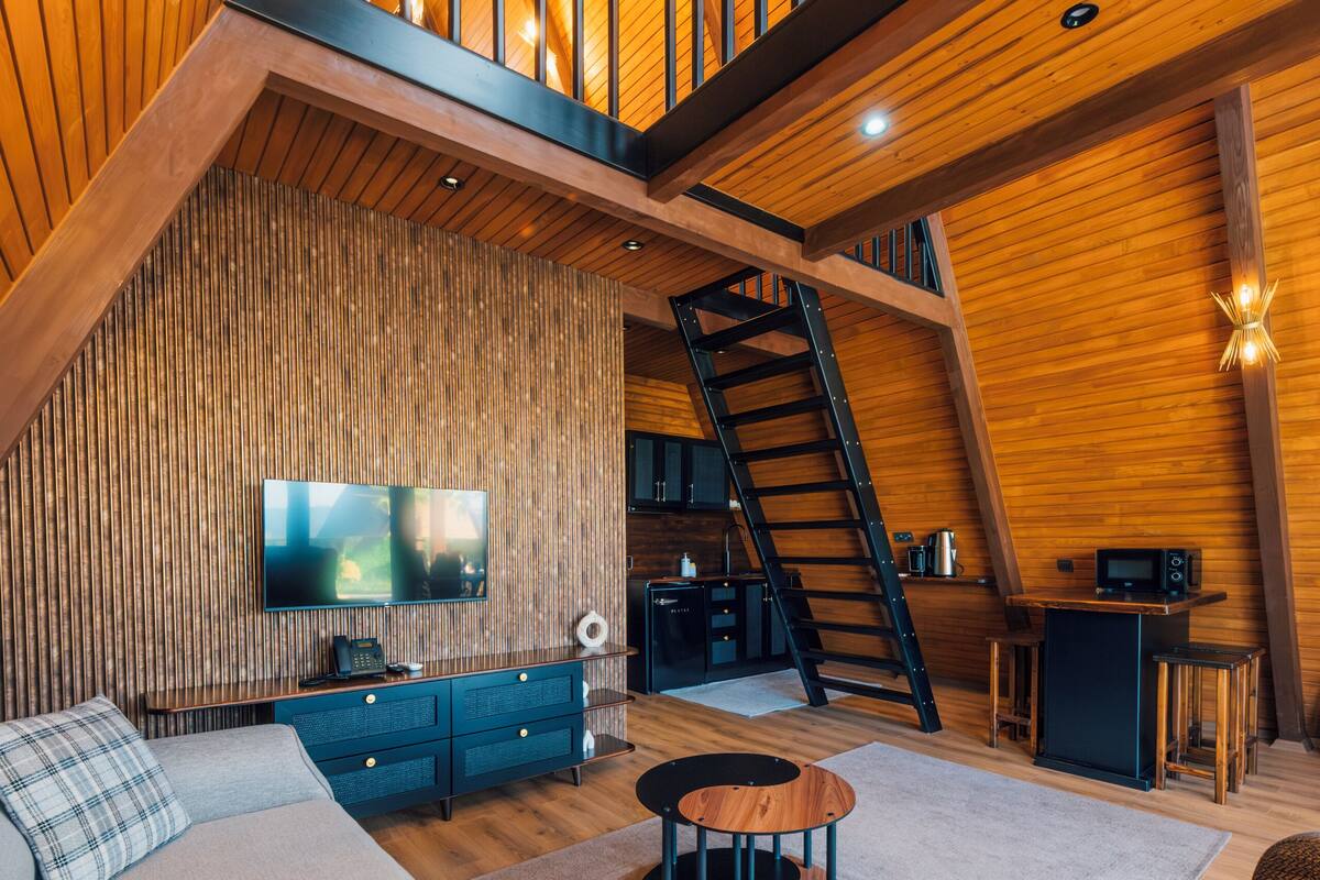 A spacious loft-style living area with a high ceiling, wooden beams, a staircase, a flat-screen TV, a sofa, a coffee table, and a kitchen area.