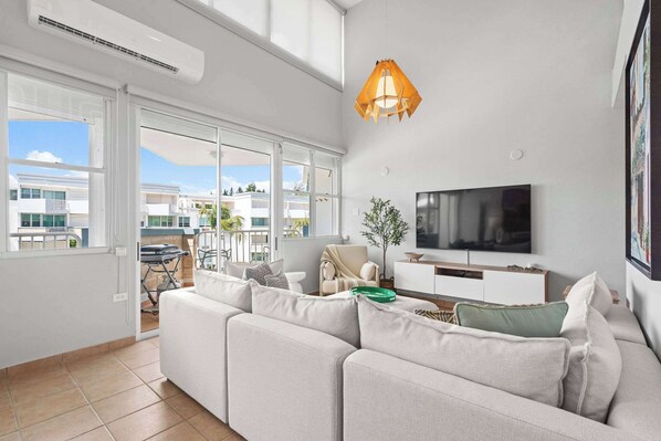 A modern living room with a large sofa, a flat-screen TV, and a dining area visible through glass doors.
