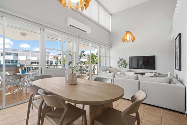 A modern living room with a large TV, a dining area with a wooden table, and a sofa.
