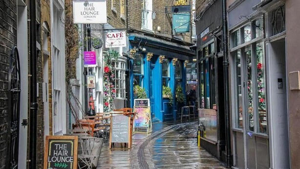 A narrow street with a blue building, signs for 'The Hair Lounge' and 'Cafe', and outdoor seating.