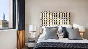 A hotel room with a bed, pillows, a nightstand, a lamp, and a view of a building through the window.
