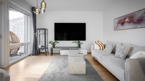 Un soggiorno moderno con una grande TV a schermo piatto, un divano grigio, un tavolino da caffè e un'ampia finestra che si affaccia su edifici.