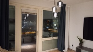 Un soggiorno moderno con una porta scorrevole in vetro che dà su un balcone, una tenda blu scuro, un lampadario a soffitto e una piccola pianta su un tavolo consolle.