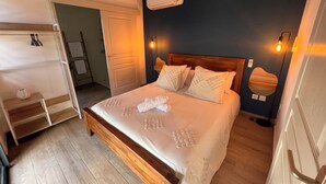 A neatly made bed with a wooden headboard and white linens, a bedside table with a lamp, and a wall-mounted air conditioner. - Longanea Residence Entière (Saint Pierre)