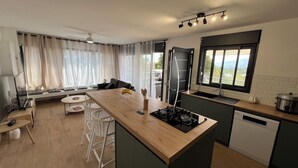 A modern kitchen with a central island, a dining area with white chairs, and a view of greenery through the window. - Longanea Residence Entière (Saint Pierre)