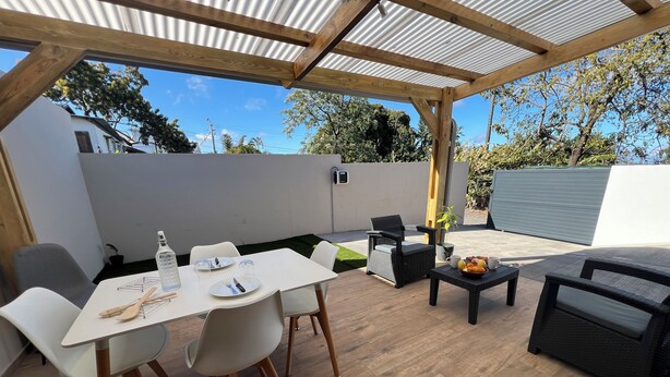 An outdoor patio with a wooden table and chairs, a small table with fruits and cups, and a pergola overhead. - Longanea Residence Entière (Saint Pierre)