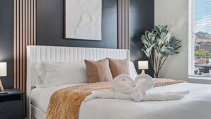 Ein ordentlich bezogenes Bett mit einem weißen Kopfteil, einer braunen Decke und zwei Kissen. Darüber hängt ein gerahmtes Kunstwerk und rechts daneben ein Blumentopf.