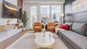 A modern living room with a sofa, two armchairs, a coffee table, and a TV mounted on the wall. There are large windows with blinds, a potted plant, and a lamp.