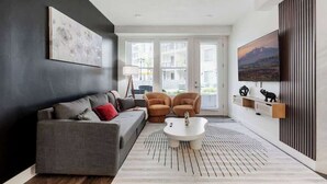 A modern living room with a grey sofa, two armchairs, a coffee table, and a large TV on the wall.
