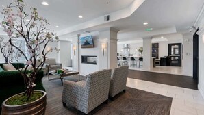 A spacious lobby with a green sofa, patterned armchairs, a large potted tree, and a modern kitchen area in the background. - Stylish 2-Bedroom with Parking, Near Temple Square (Salt Lake City)