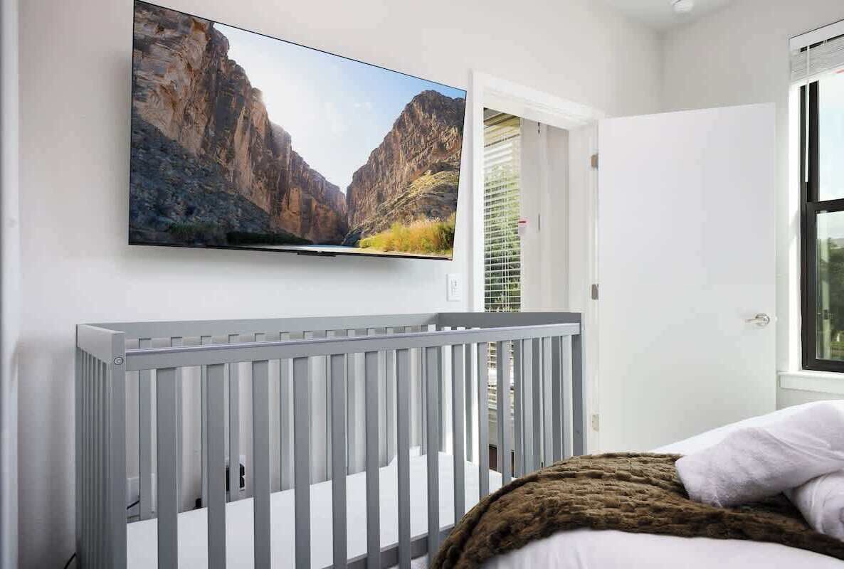 A room with a white crib, a mounted TV displaying a canyon landscape, and a window with blinds.