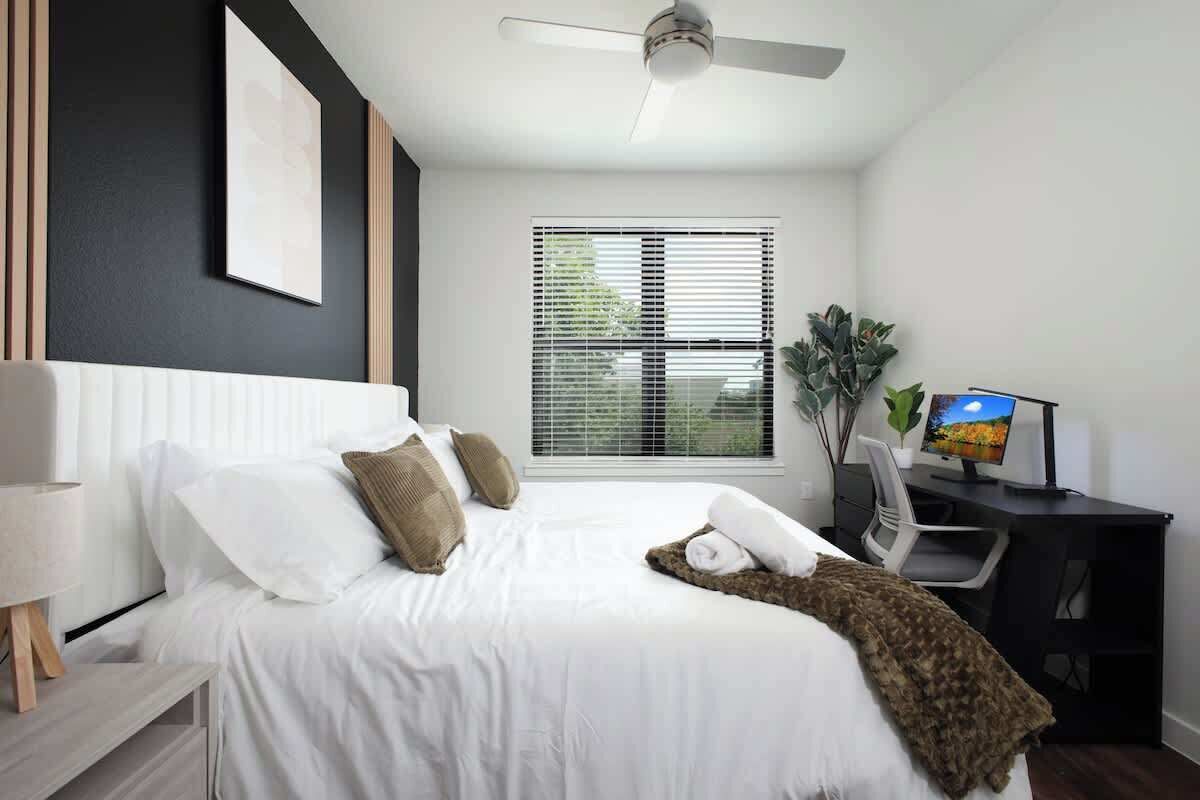 Una habitación de hotel moderna con cama, escritorio, silla, ventilador de techo y ventana con persianas.