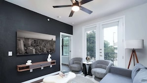 Een moderne woonkamer met een grijze bank, een witte salontafel en grijze fauteuils. Er is een plafondventilator, een groot zwart-wit kunstwerk aan de muur en een vloerlamp.