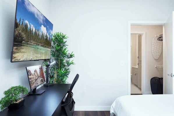 Habitación de hotel con cama, escritorio con monitor de computadora, televisión y planta en maceta.