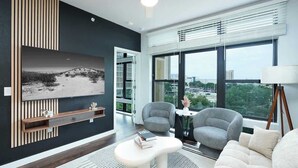 Een moderne woonkamer met een grote televisie, twee grijze fauteuils, een witte bank en een houten plank met decoratieve objecten.