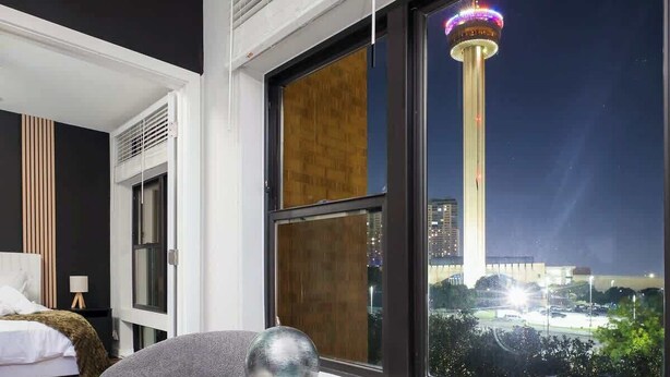 A modern hotel room with a large window offering a nighttime city view, a round table with a spherical object, and a cozy seating area. - Snug 1-Bedroom with Gym,  Near Riverwalk & Dome (San Antonio)