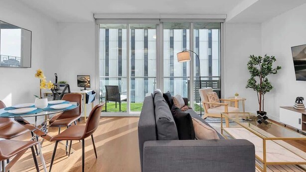 A modern living room with a sofa, dining table, and chairs. There is a large window with a view of the outside.