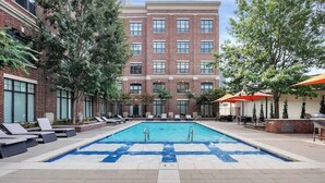 A rectangular pool with lounge chairs and umbrellas, surrounded by brick buildings and trees.