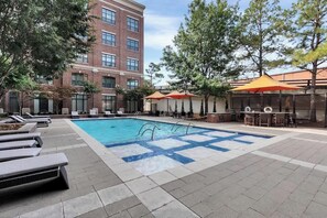 Une piscine entourée d’arbres et d’un bâtiment en briques, avec des chaises longues et des parasols à proximité.