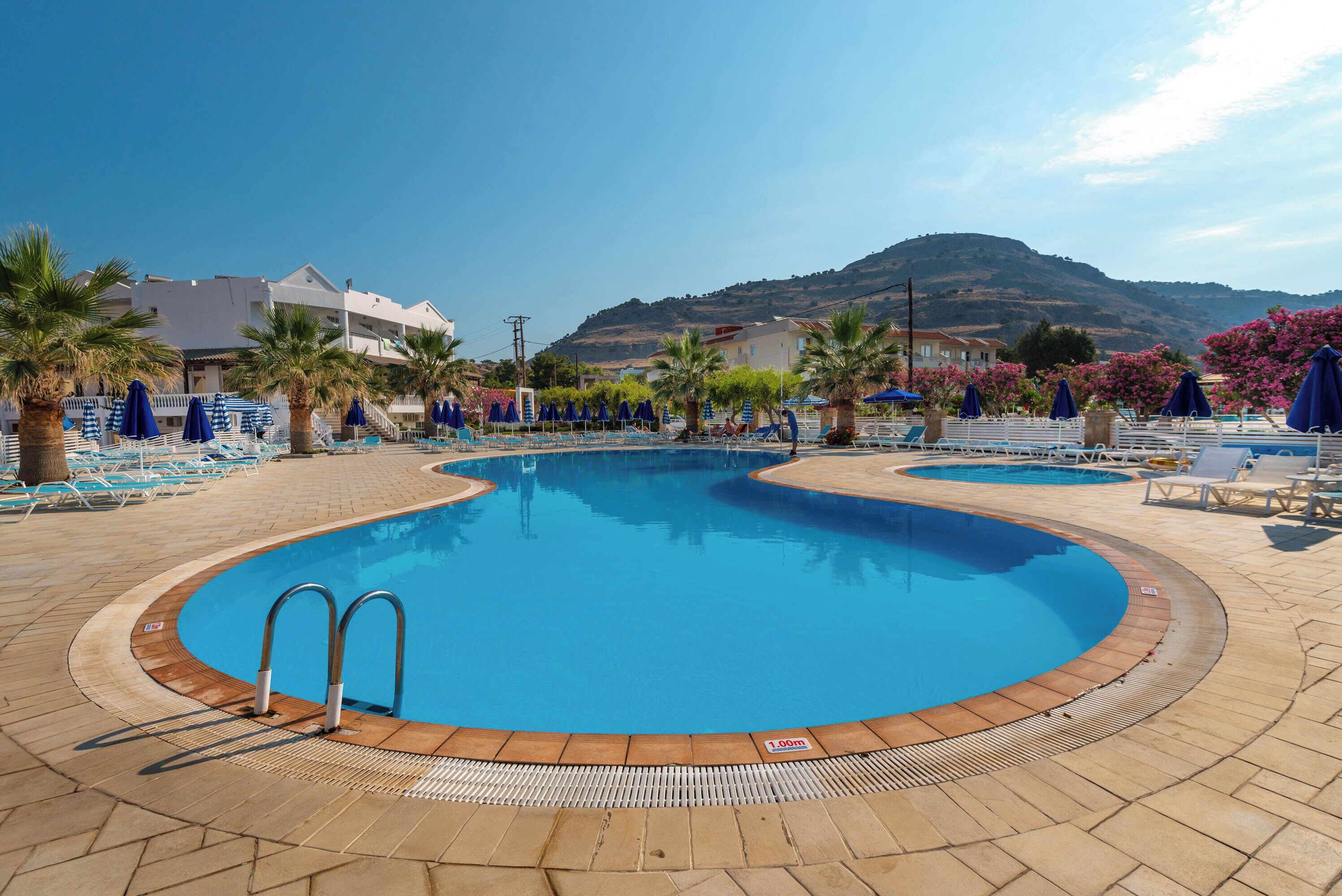 Une piscine entourée de palmiers et d’immeubles, avec des chaises longues et des parasols.