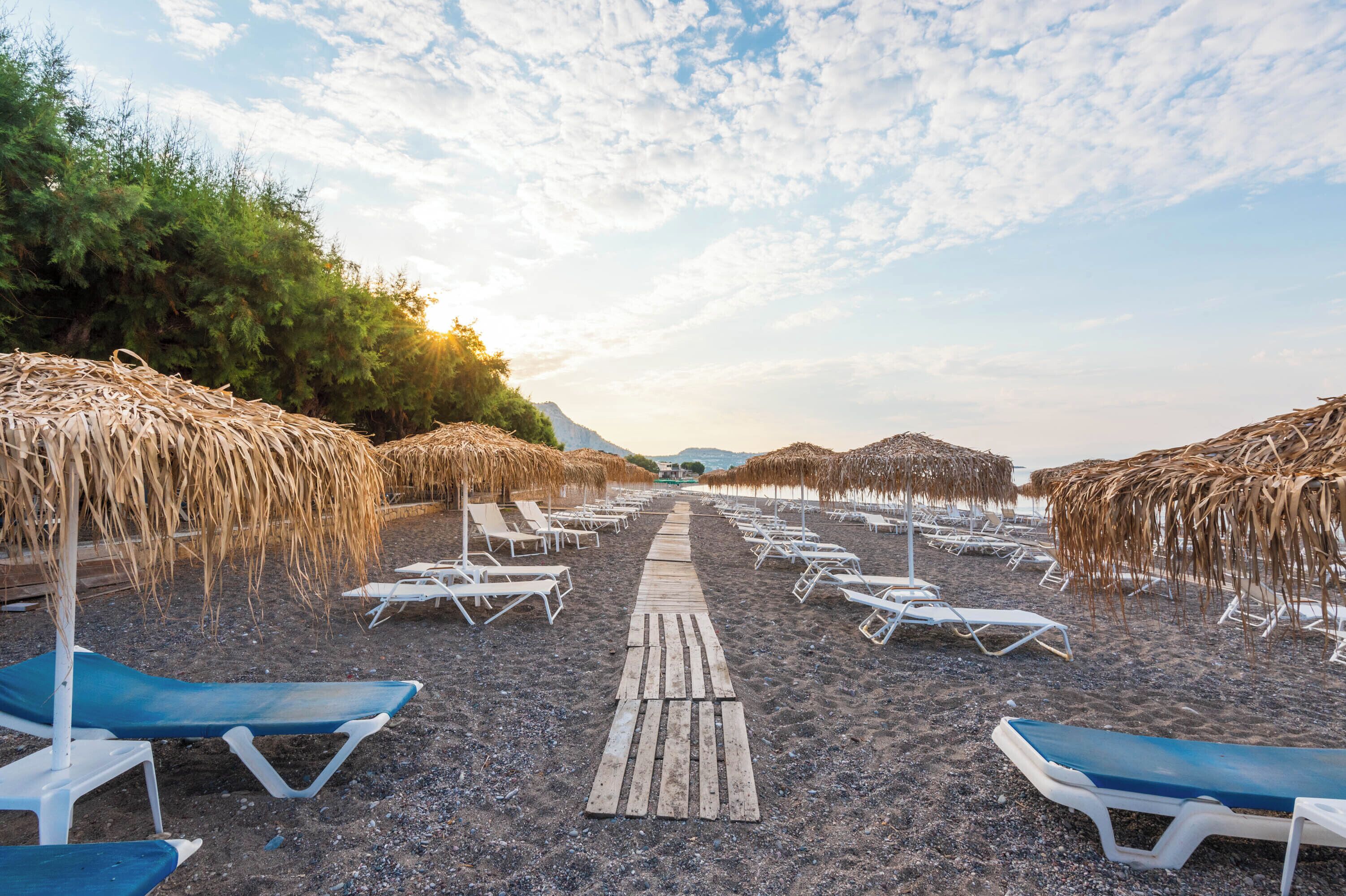 Une plage bordée de rangées de chaises longues et d’ombrelles en chaume, une promenade en bois et un ciel dégagé.