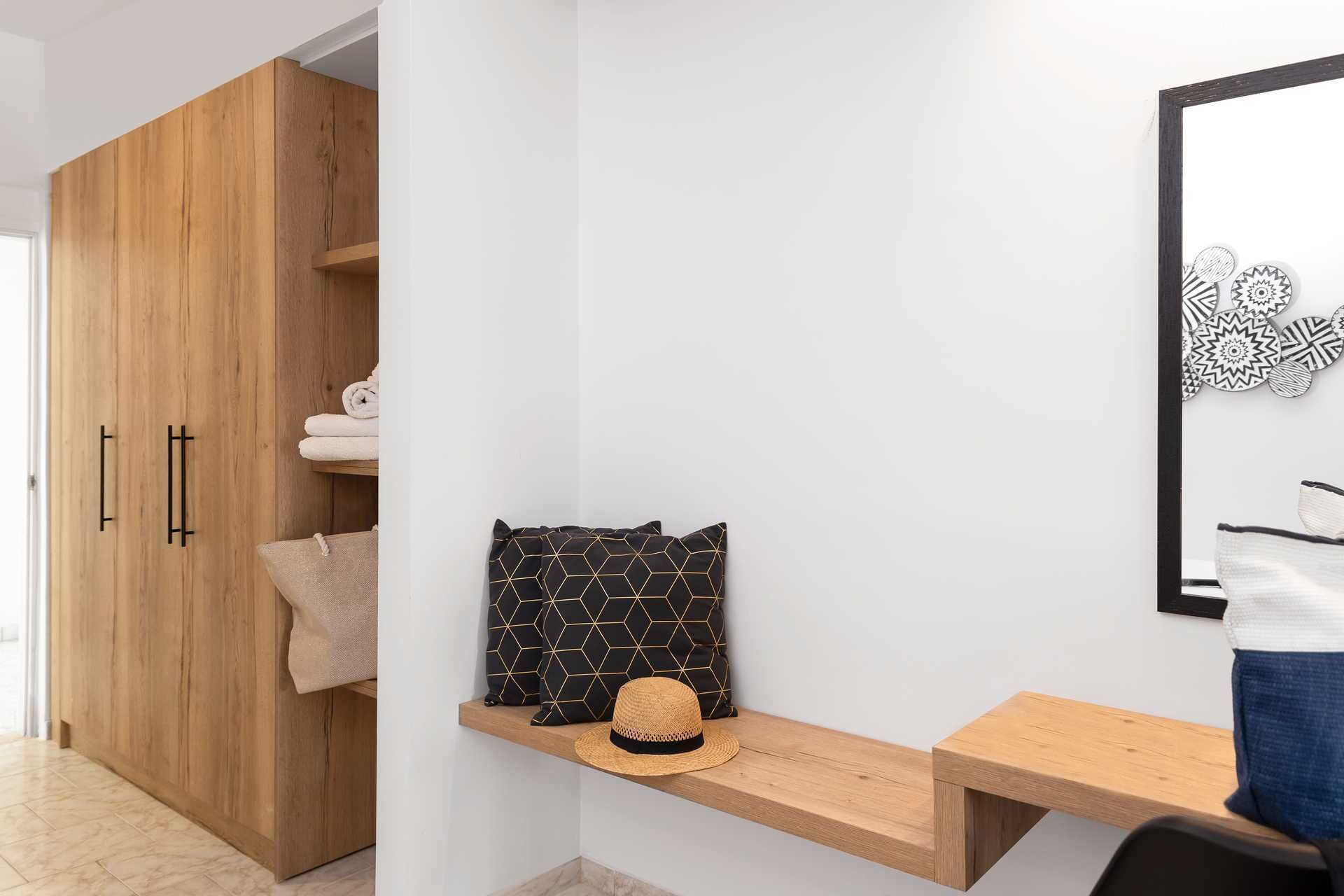 Een kamer met een houten garderobe, een plank met handdoeken, een zwart-en-goud gestreept kussen, een strooien hoed en een houten bank.