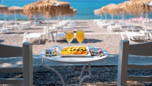 Una mesa con un plato de comida y dos vasos de una bebida amarilla, sobre una playa con sombrillas y sillas de descanso.