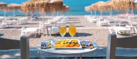 A table with a plate of food and two glasses of a yellow beverage, set on a beach with umbrellas and lounge chairs.