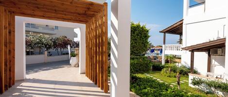 Un corredor exterior moderno con vigas de madera, paredes blancas y vista a una piscina y vegetación.
