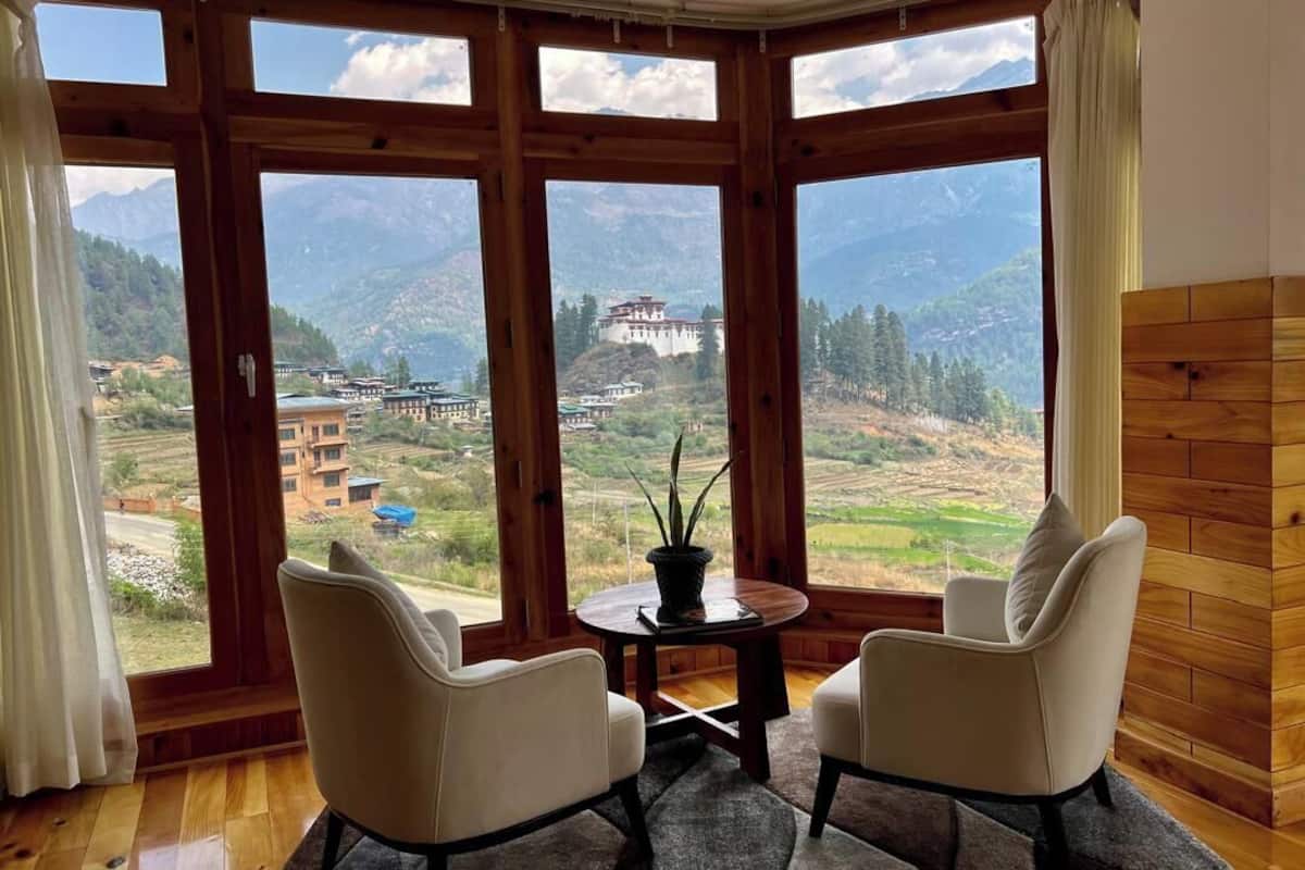 A room with large windows offering a view of a mountainous landscape and a traditional building. There are two chairs and a small table with a plant.