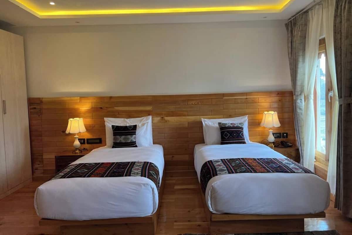 A hotel room with two beds, wooden headboard, bedside lamps, and a window with curtains.