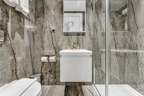 Un baño moderno con salpicadero de mármol, un lavabo blanco con dos grifos, un inodoro y una cabina de ducha con mampara de vidrio.
