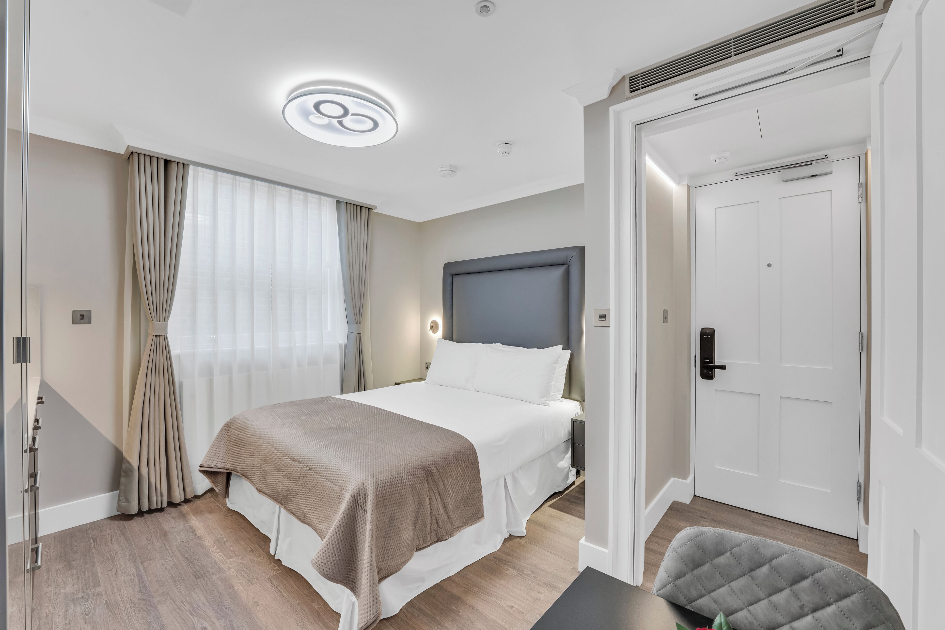 A modern hotel room with a large bed, a bedside table, a chair, and a door.