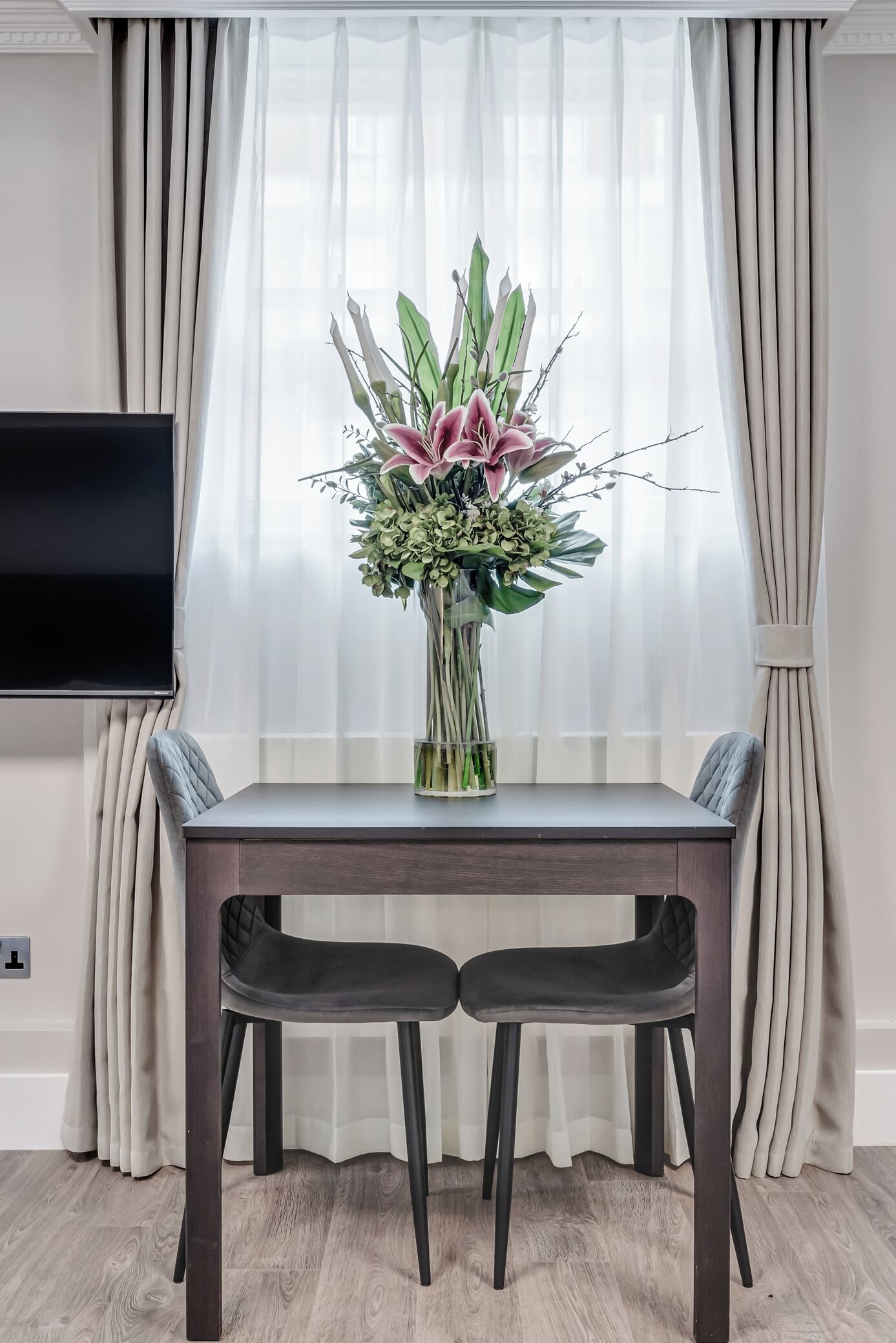 A dining area with a table, two chairs, a vase of flowers, and a TV on the wall.