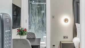 Una moderna camera d'albergo con pavimento in legno, una scrivania con telefono, una sedia, un piccolo comodino con lampada e un letto con testiera.