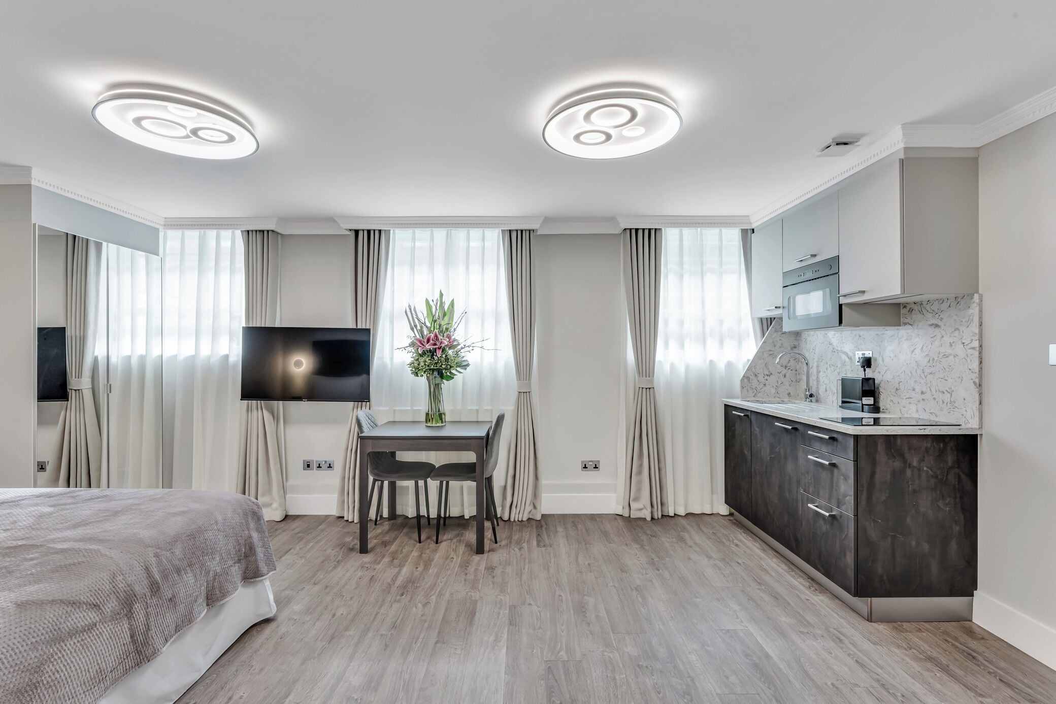 Ein modernes Hotelzimmer mit einem Bett, einem Esstisch mit Stühlen, einem Fernseher, einer Küchenzeile mit Schränken und einer Deckenleuchte.
